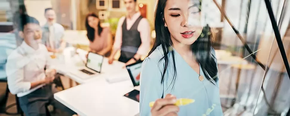 Woman writing on a glass board during a team meeting in an office.