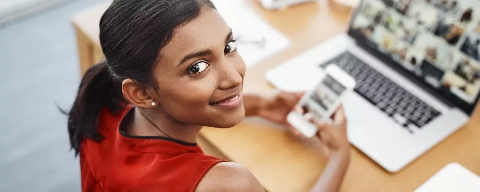 Woman in red top using smartphone and laptop at desk, smiling.