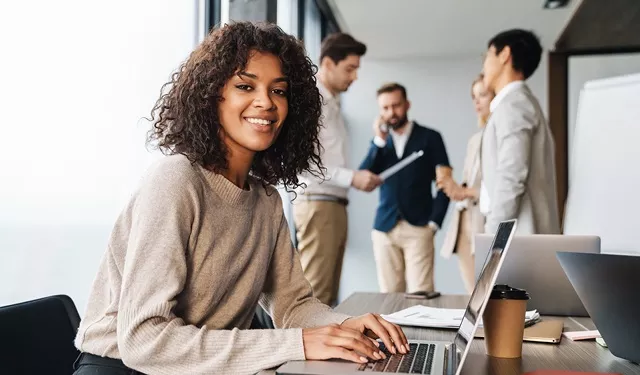 A smiling person works on a laptop at a desk, while colleagues have a discussion in the background.