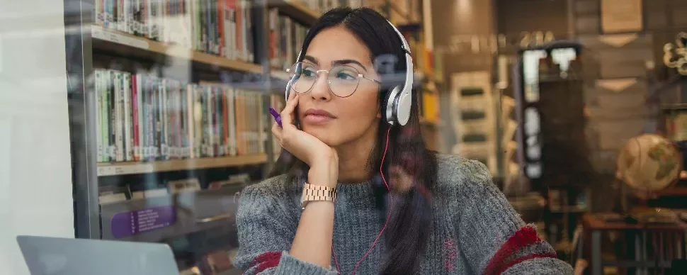 Woman with headphones studies at a library, gazing pensively outside.