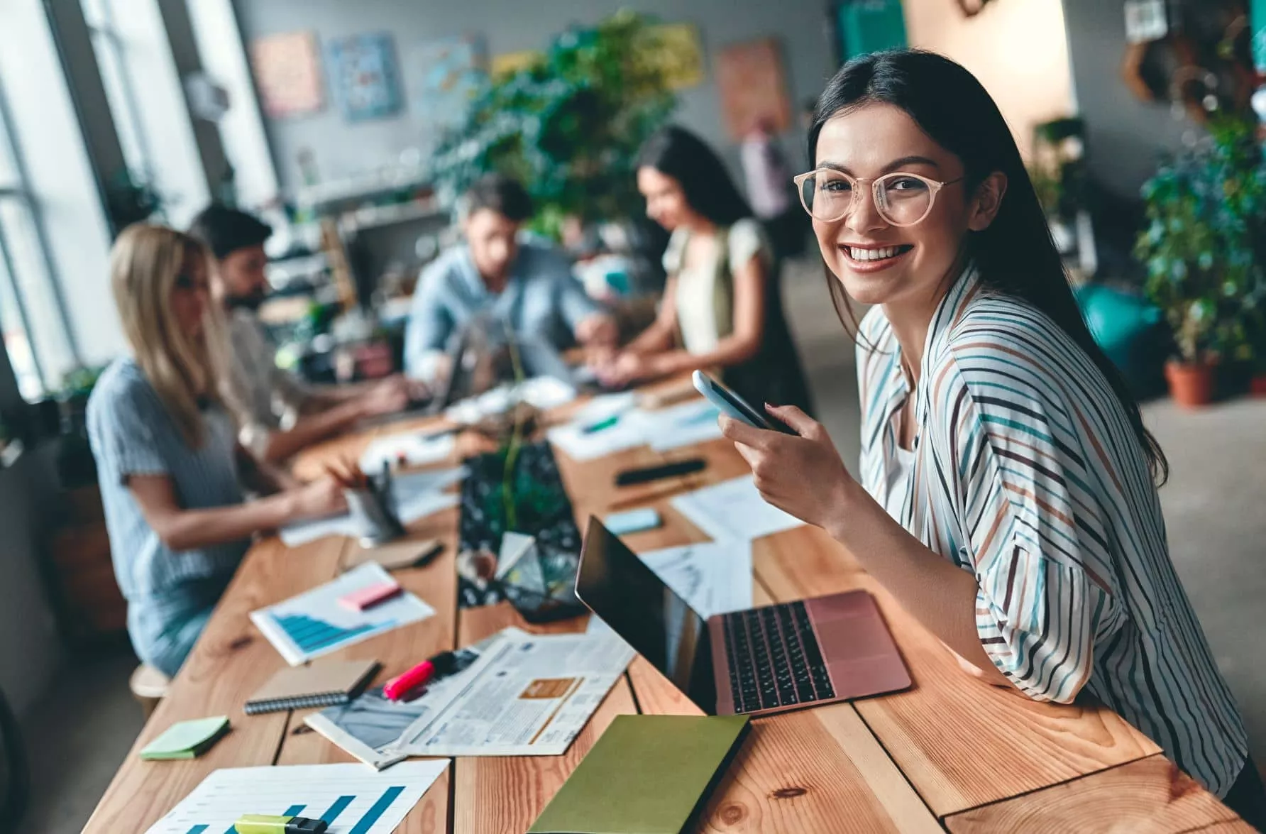Smiling woman in glasses with coworkers in a casual office setting.