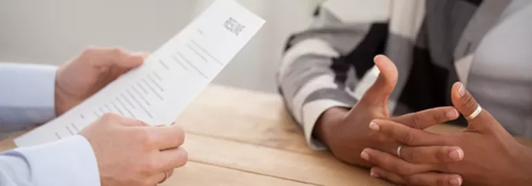 Close-up of an interview, one person holding a resume, another with folded hands on a wooden table.