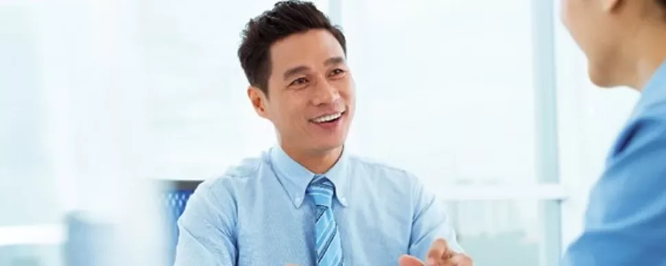 Smiling man in a blue shirt talking to a colleague in an office.