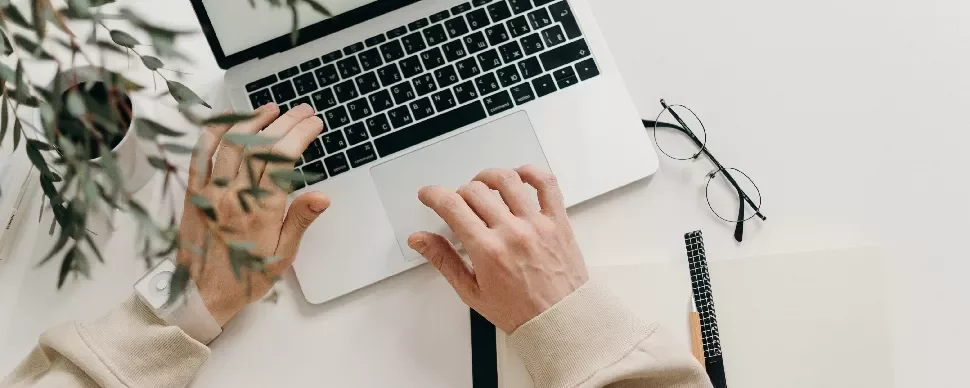 Person typing on a laptop with a notebook, pen, and glasses on a white desk.