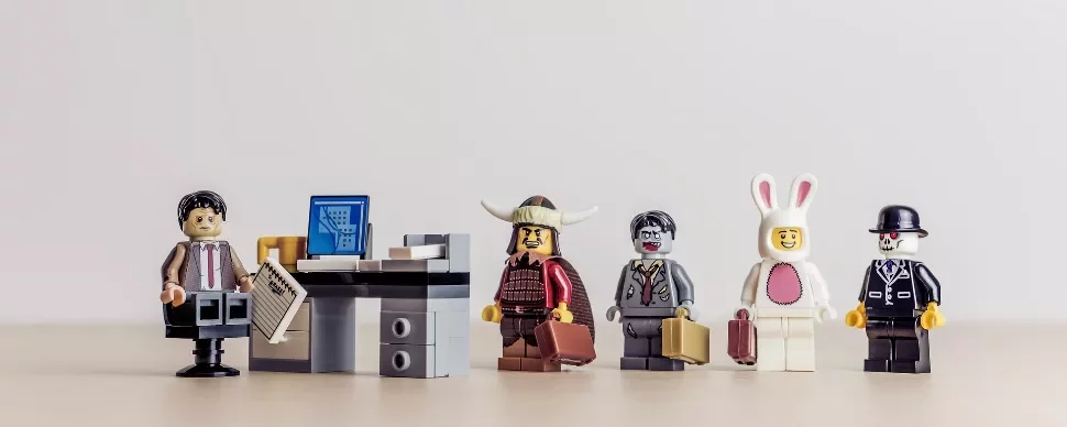 LEGO figures in costumes stand in a line, one sits at a desk with a paper, depicting a quirky office scene.