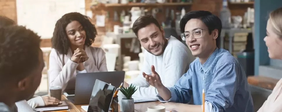 Diverse group of people in a casual meeting, discussing and smiling around a table with laptops.