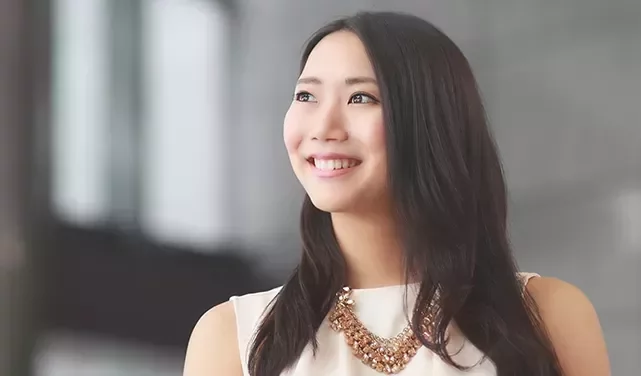 Smiling woman with long hair and necklace against a blurred background.