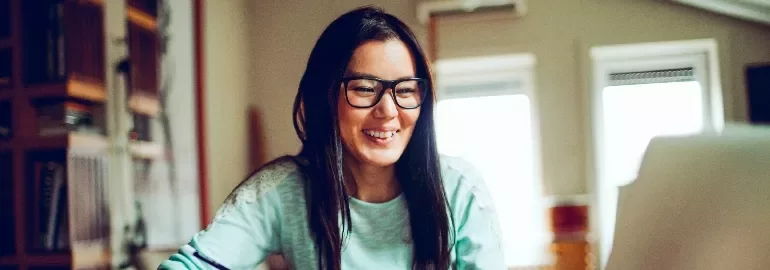 A woman with glasses smiling while working on a laptop at home.