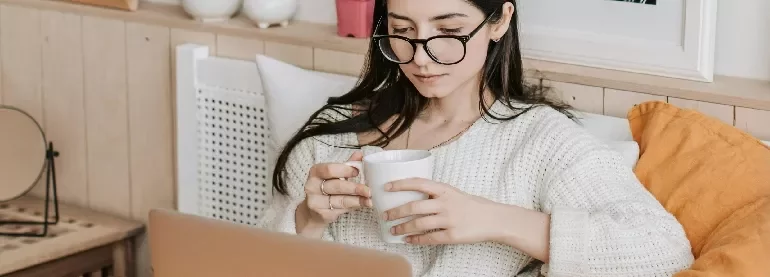 Woman wearing glasses, holding a mug and looking at a laptop.